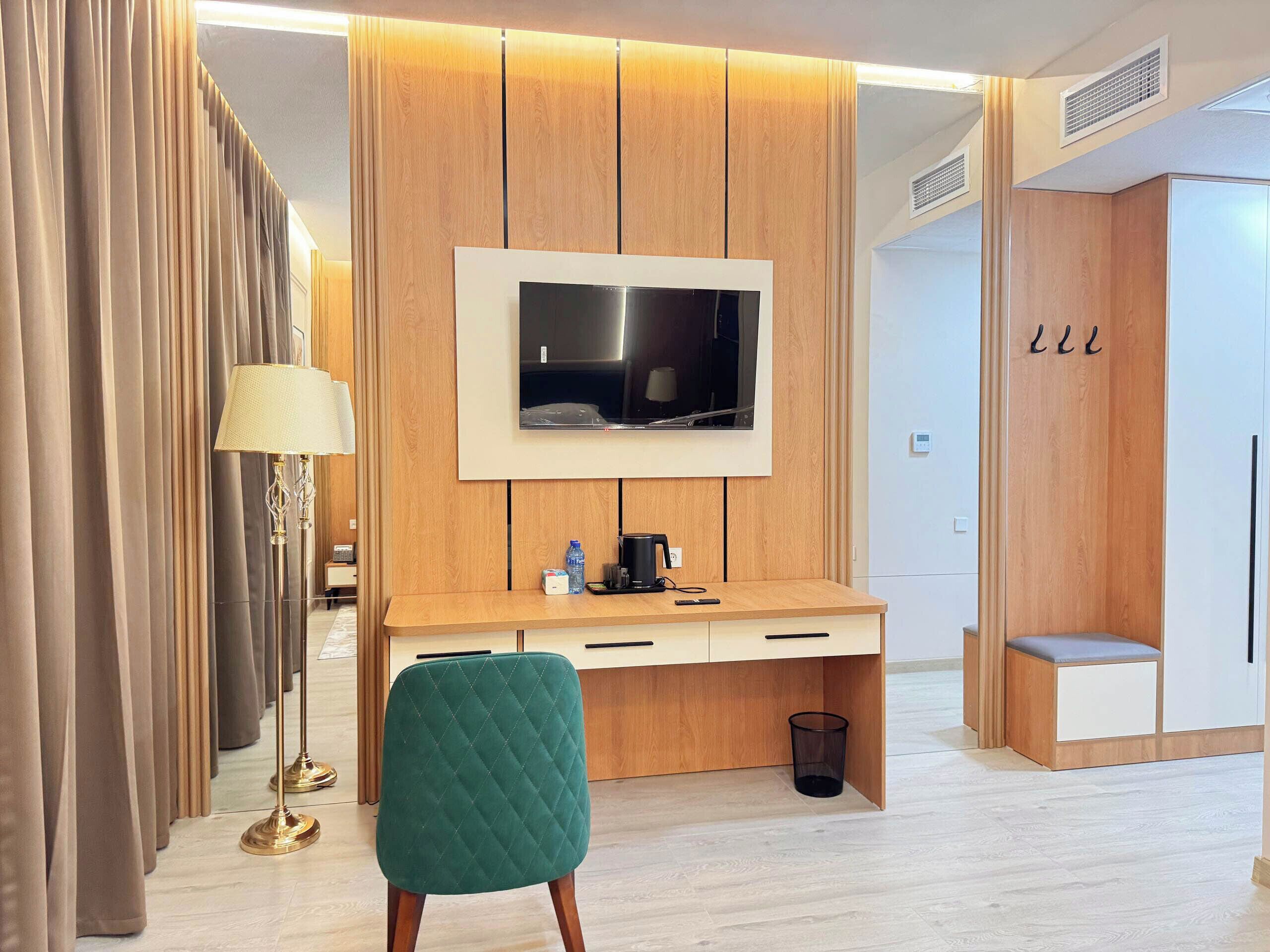 A modern hotel room with a wooden desk, a television mounted above it, a green chair, a floor lamp, and a wardrobe with a mirror.
