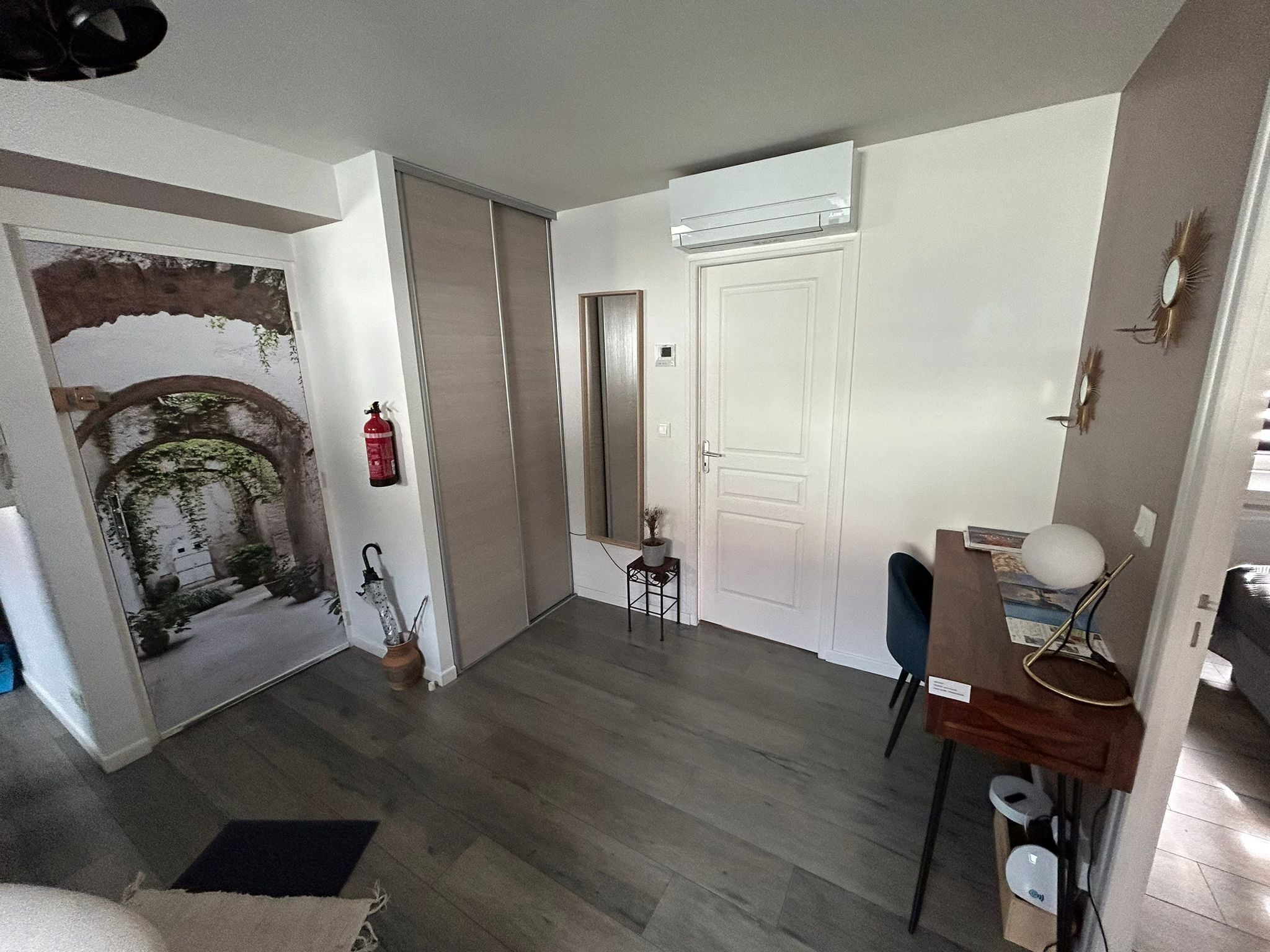 A room with a wooden floor, a white door, a small table with a chair, a wall-mounted air conditioner, and a decorative archway.