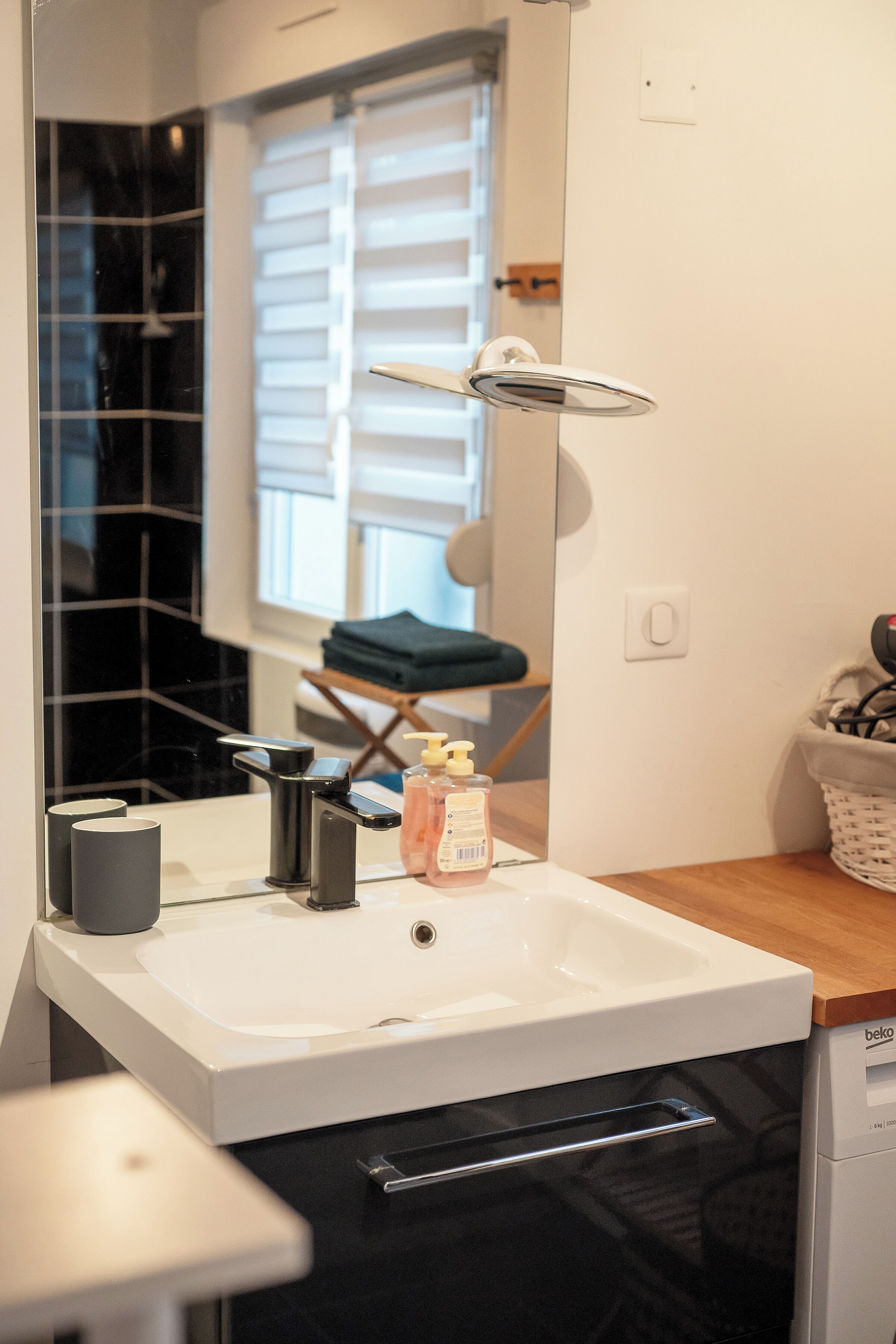 Een badkamer met een witte wastafel, een spiegel, een zwarte handdoek en een raam met zonwering.