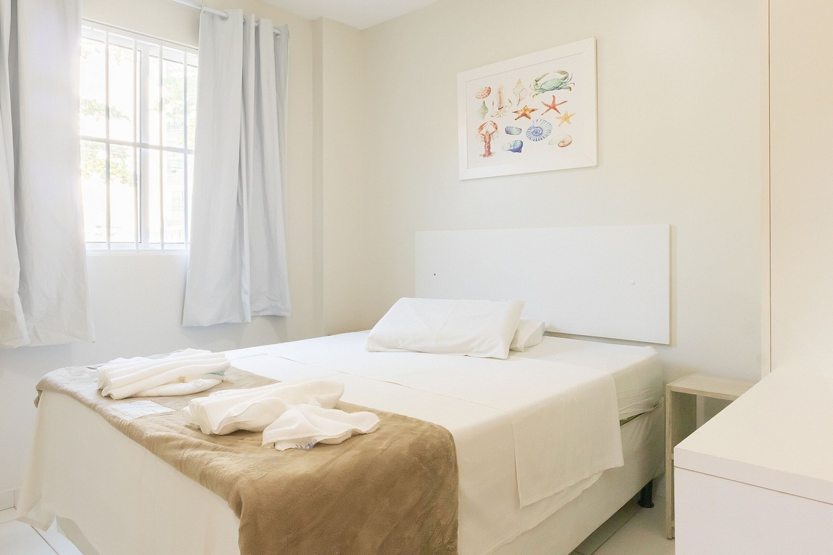 Ein Schlafzimmer mit Bett, weißen Vorhängen, einem Nachttisch, einem kleinen Beistelltisch und einem Wandbild.