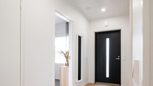 Un pasillo con paredes blancas, una puerta negra y una ventana con un jarrón de plantas secas.