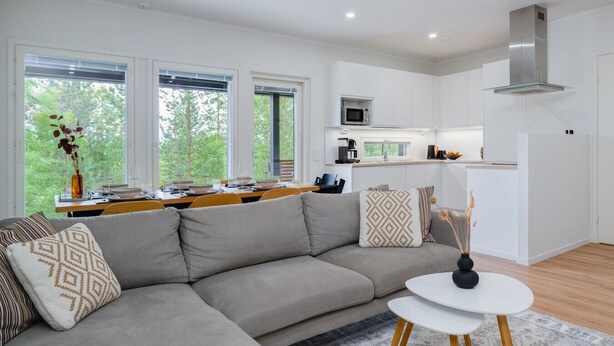 Una sala de estar moderna con un sofá modular, una mesa de comedor con sillas, una zona de cocina con armarios blancos y ventanas amplias con vistas a árboles.