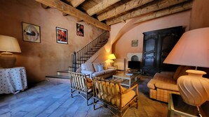 A rustic interior with exposed wooden beams, a staircase, a sofa, a wicker chair, a television, and a lamp.