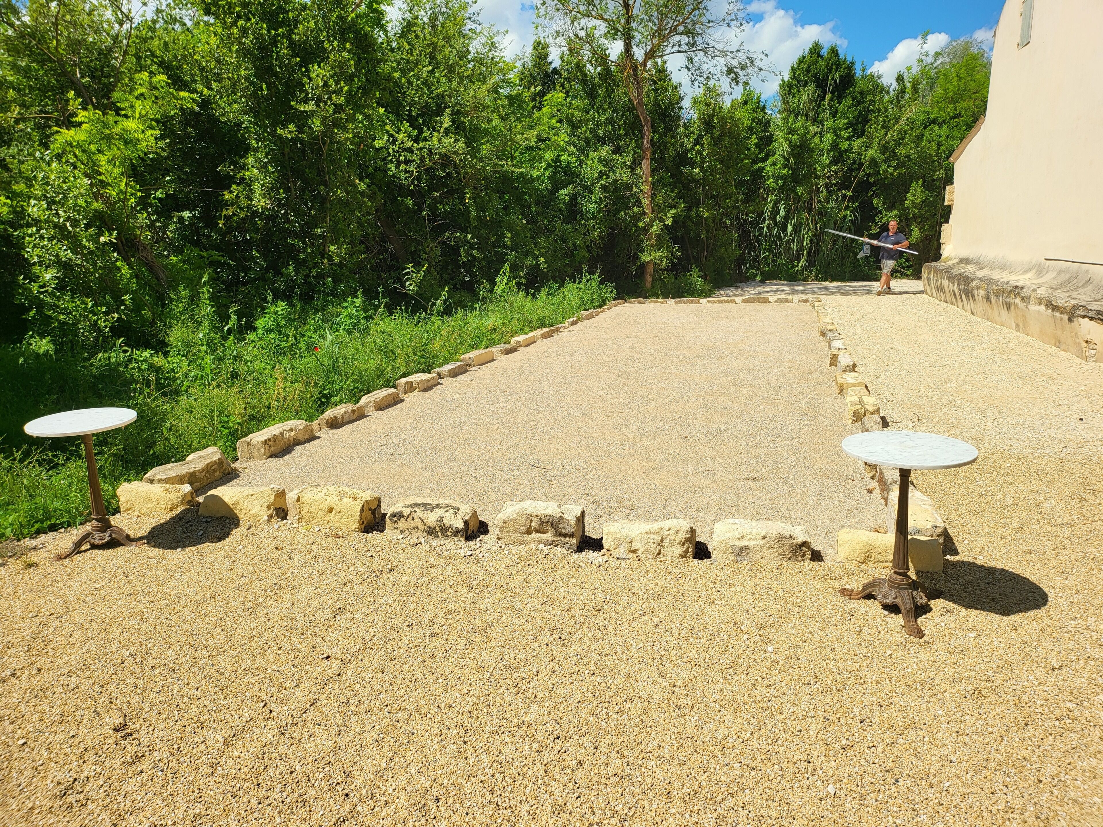 A gravel area with two round tables, surrounded by a low stone wall and dense greenery.