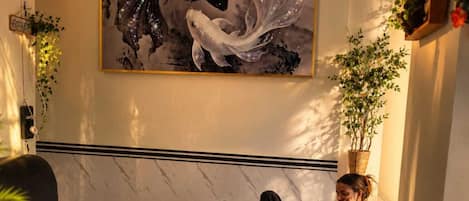 A room with a marble floor, a wooden coffee table with books and a water bottle, a black sofa, a green chair, and a large abstract painting on the wall.