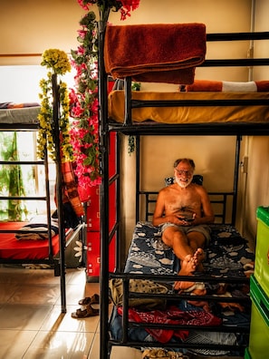 A bunk bed with a person sitting on the lower bunk, a window with curtains, and a wall-mounted shelf with a red box. - Dazhong Backpacker's Hostel (Medan)