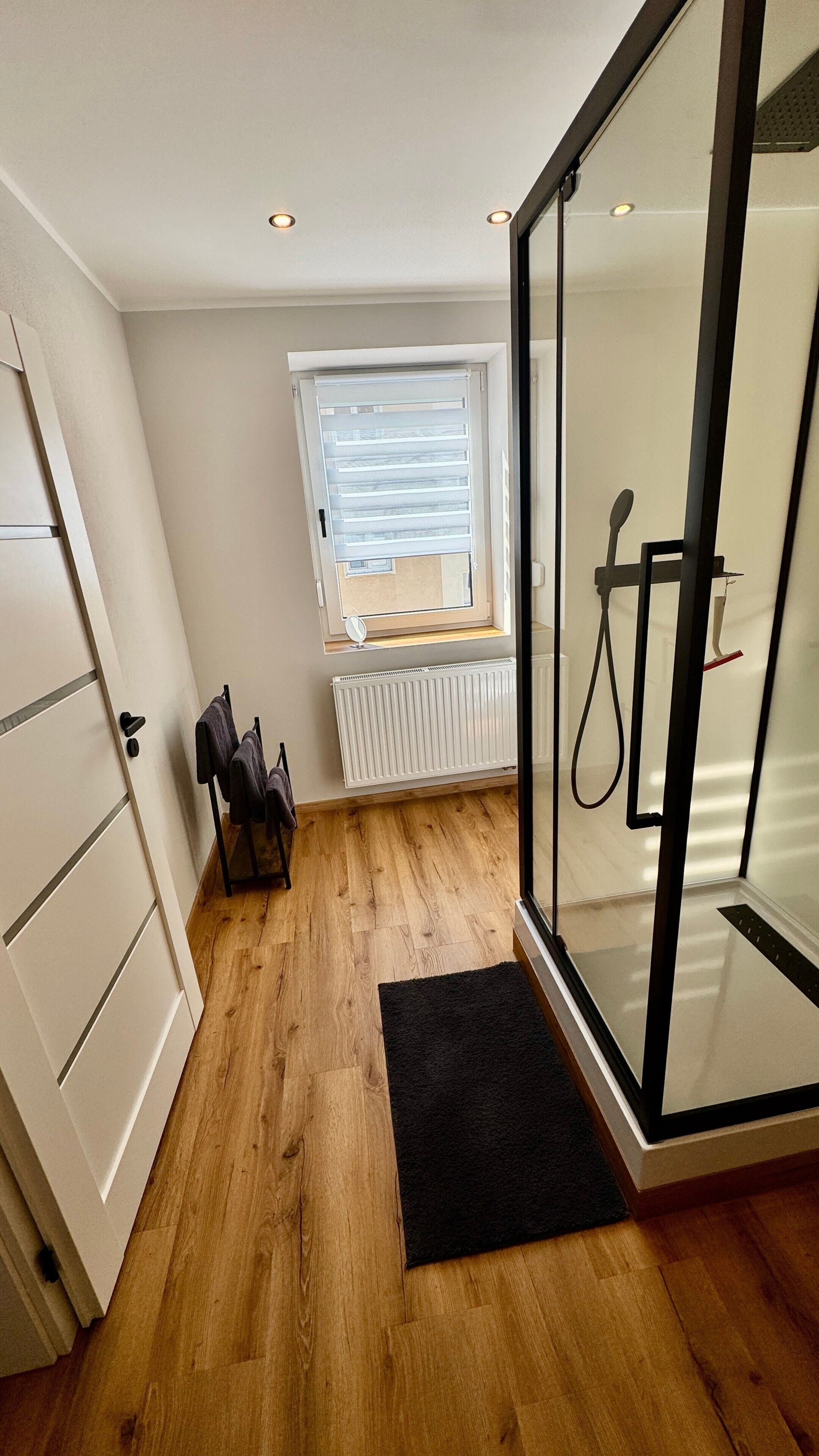 A modern bathroom with a shower, a radiator, and a window with blinds.