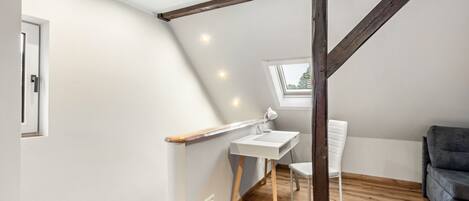 A small, well-lit attic bedroom with a sloped ceiling, a desk, a chair, and a window.