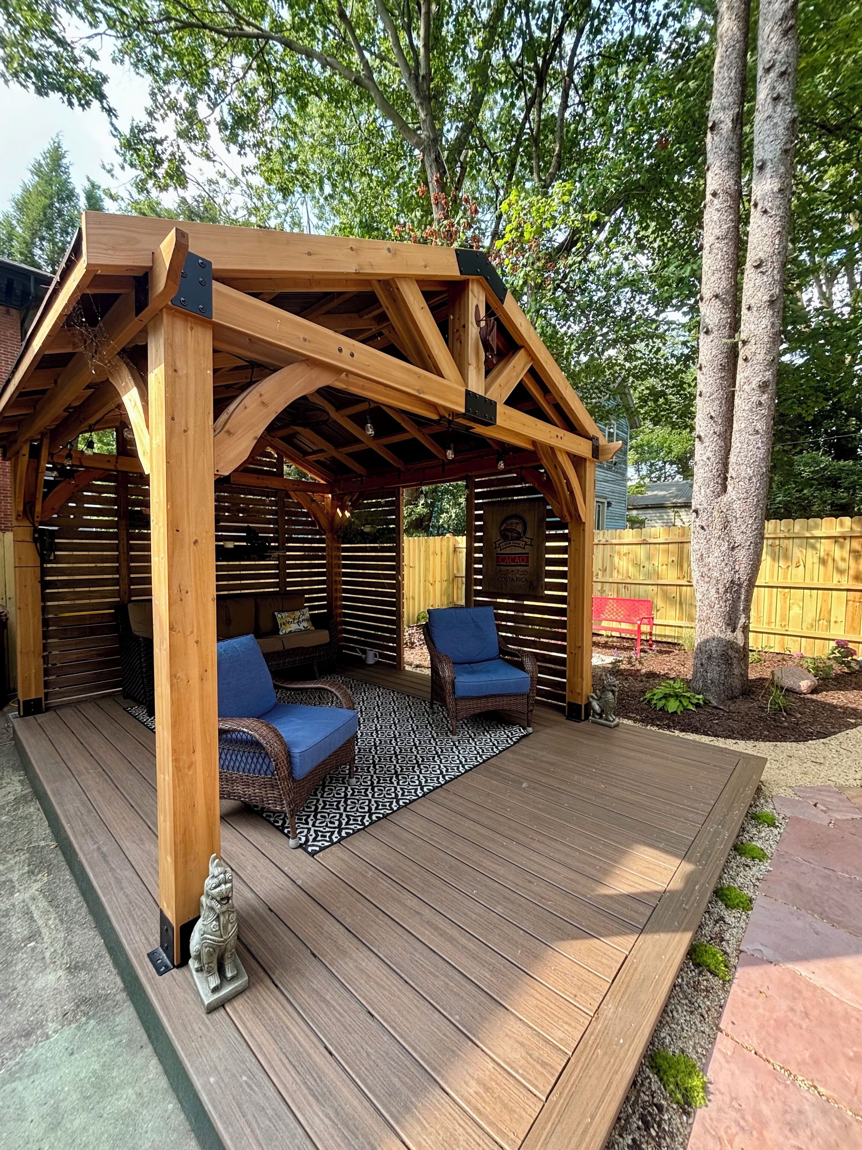 Una terraza de madera con pérgola, dos sillas de mimbre y un tapiz estampado.