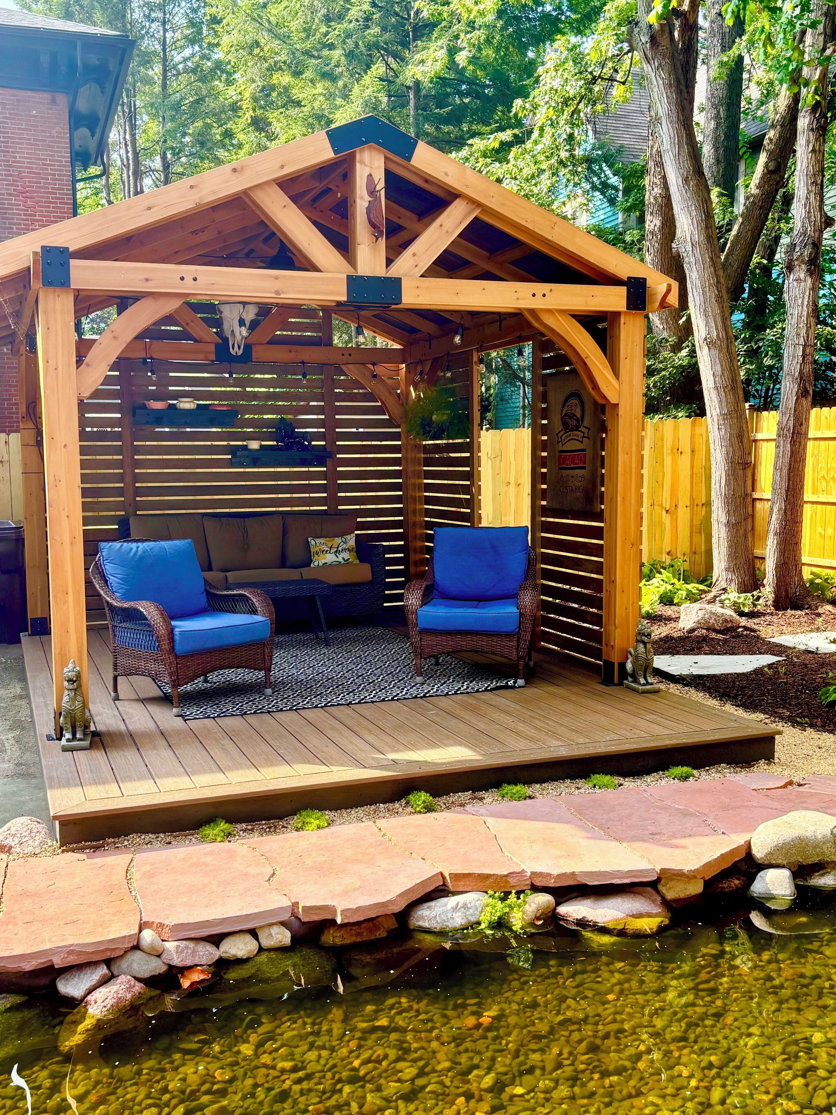 A wooden pergola with a seating area, a pond, and a wooden deck.
