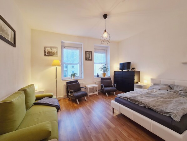 Un dormitorio moderno con cama, sofá verde, escritorio con silla, televisión y ventana con vistas a edificios.