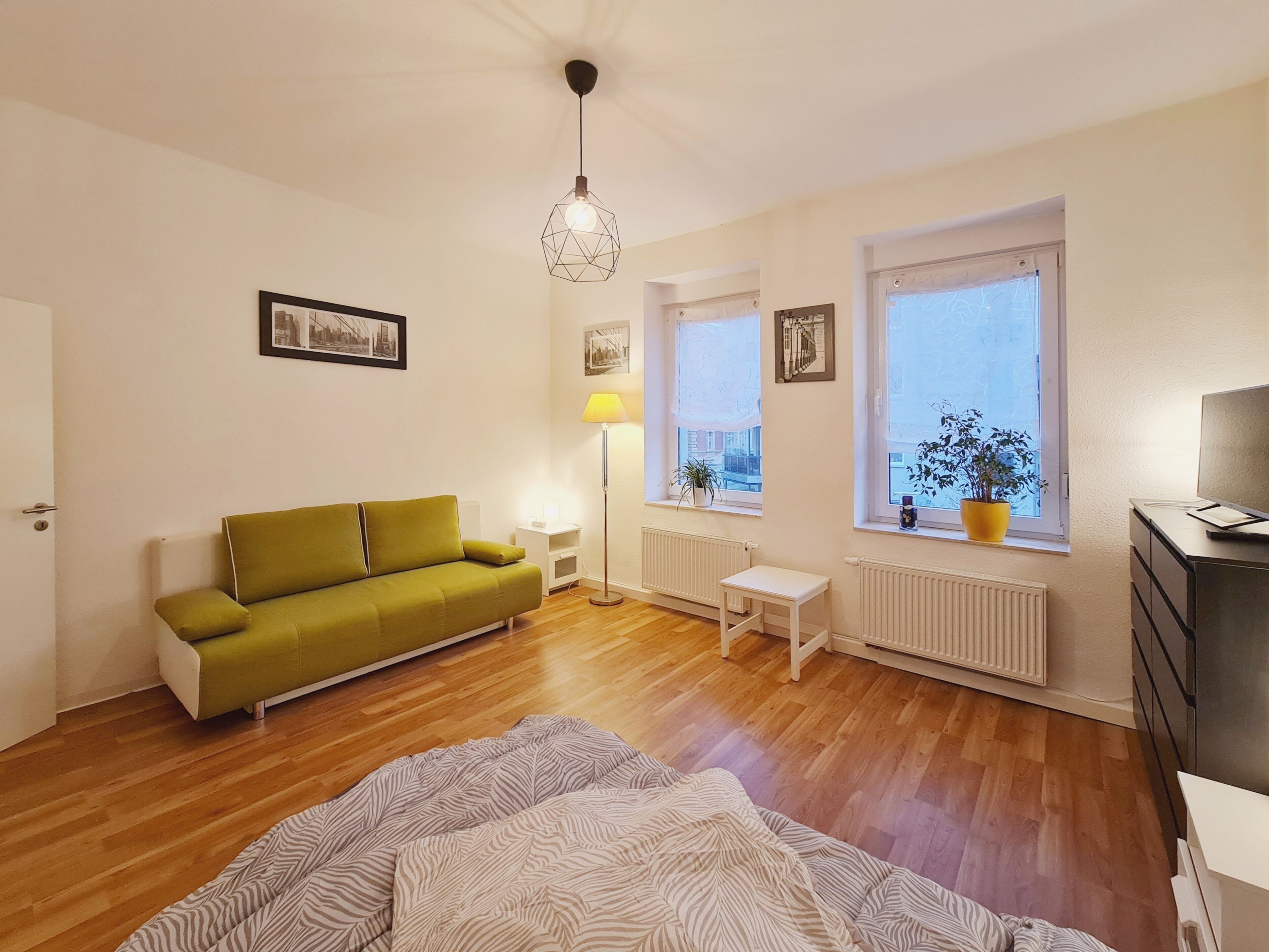 Ett rum med en grön soffa, ett litet vitt bord, en golvlampa och en säng med ett randigt täcke.