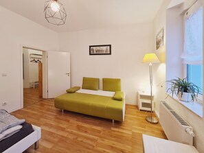 Un dormitorio con cama, mesita de noche, lámpara, radiador, ventana con planta y puerta que da a otra habitación.