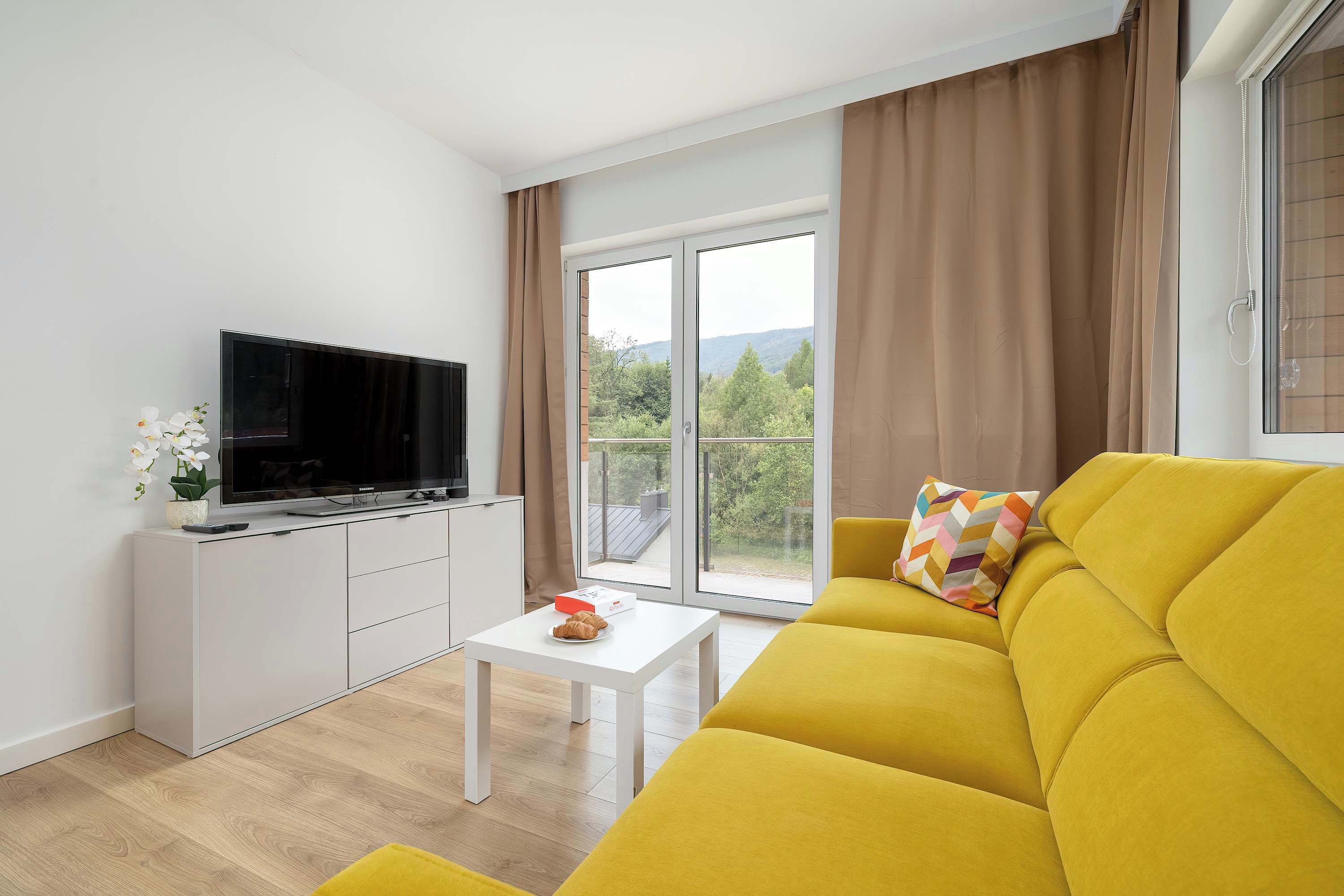 Ein modernes Wohnzimmer mit einem Flachbildfernseher auf einem weißen Schrank, einem gelben Sofa, einem kleinen weißen Tisch und einem großen Fenster mit Vorhängen.