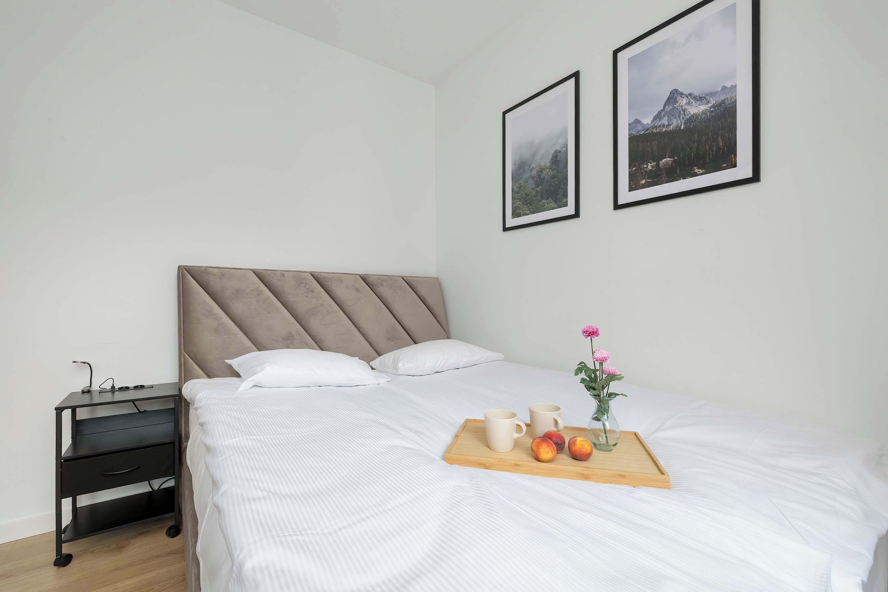 Een net opgemaakt bed met witte lakens, een nachtkastje met een lamp en een dienblad met fruit en een bloevas.