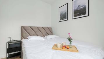 Een net opgemaakt bed met witte lakens, een nachtkastje met een lamp en een dienblad met fruit en een bloevas.