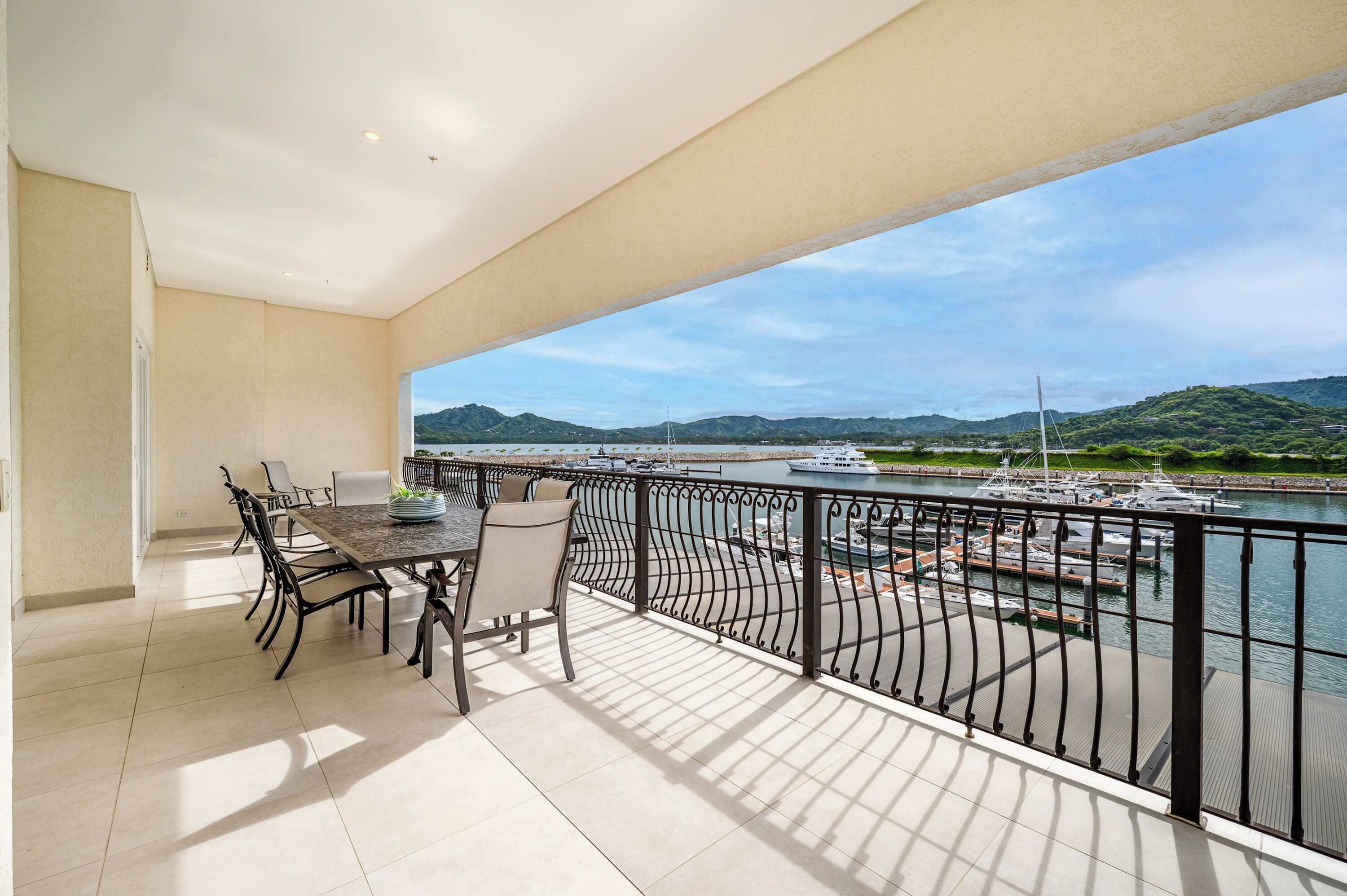 A balcony with a dining table and chairs, a view of a marina with boats, and a hilly landscape in the distance.