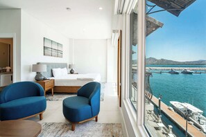Ein Hotelzimmer mit einem Bett, zwei blauen Sesseln, einem kleinen Tisch und Blick auf einen Yachthafen mit Booten.