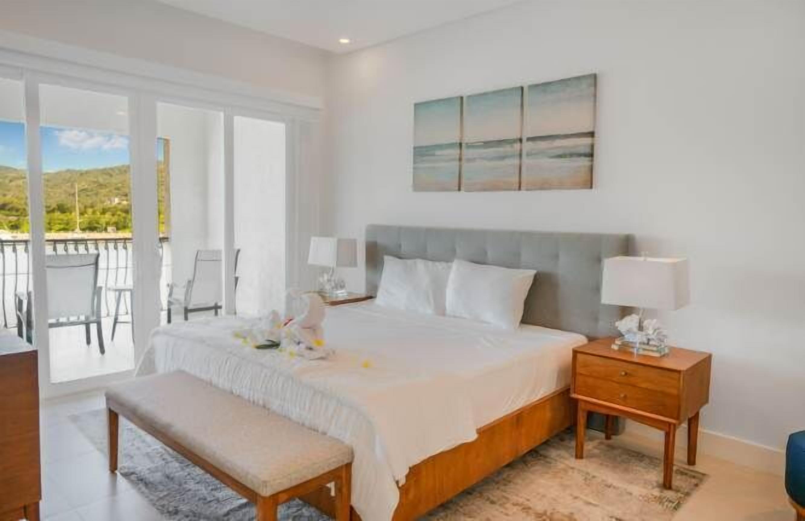 A bedroom with a bed, bedside tables, a bench, a view of the outdoors, and a painting on the wall.