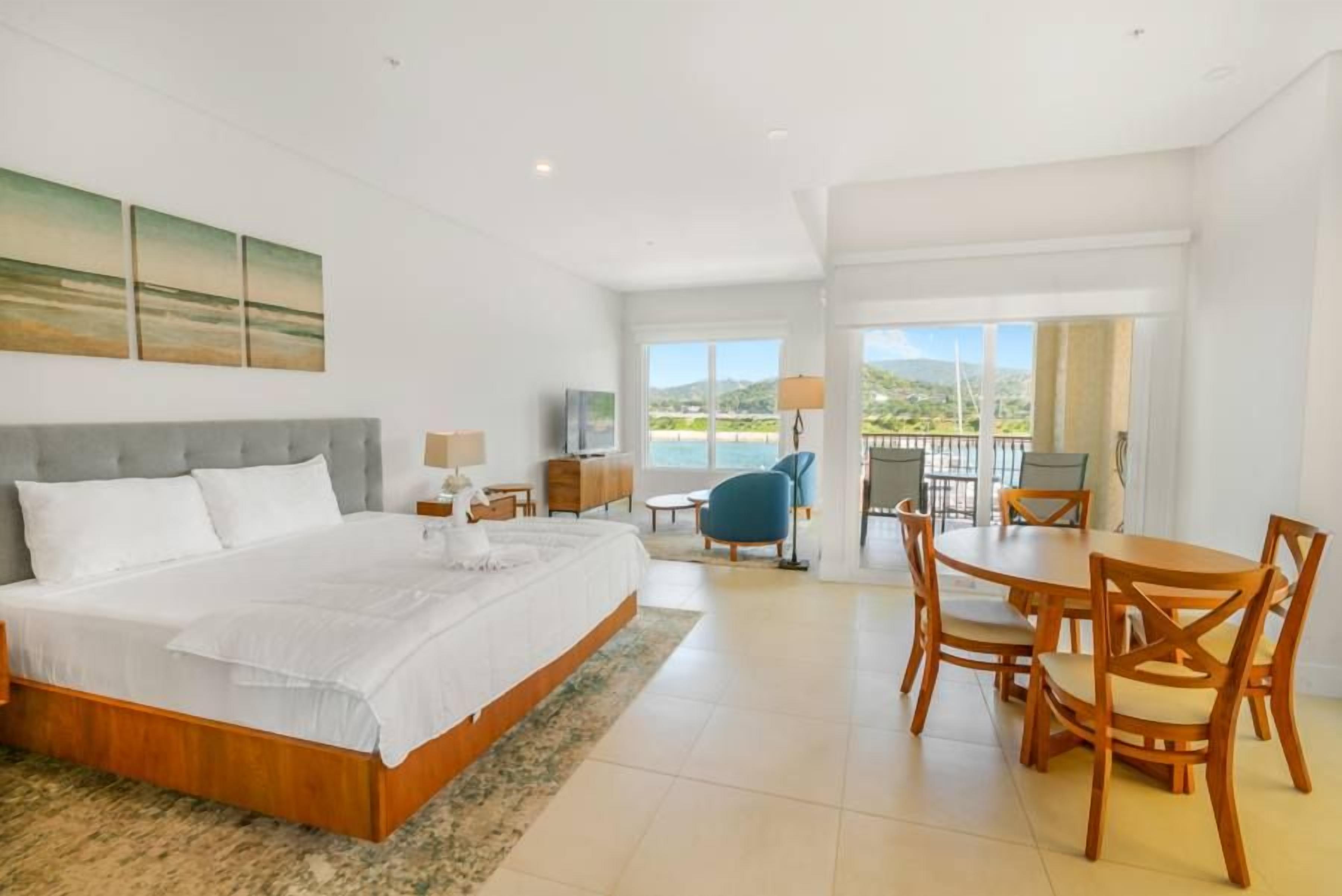 A hotel room with a large bed, a dining area with wooden furniture, and a view of the outdoors.