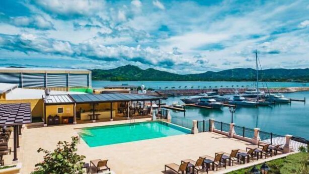 Un espace piscine avec des chaises longues, un bâtiment doté d’un toit plat et un port de plaisance avec des bateaux.