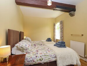 A bedroom with a bed, a bedside lamp, a radiator, and a window with curtains. - Linhay Barn (Saltash)