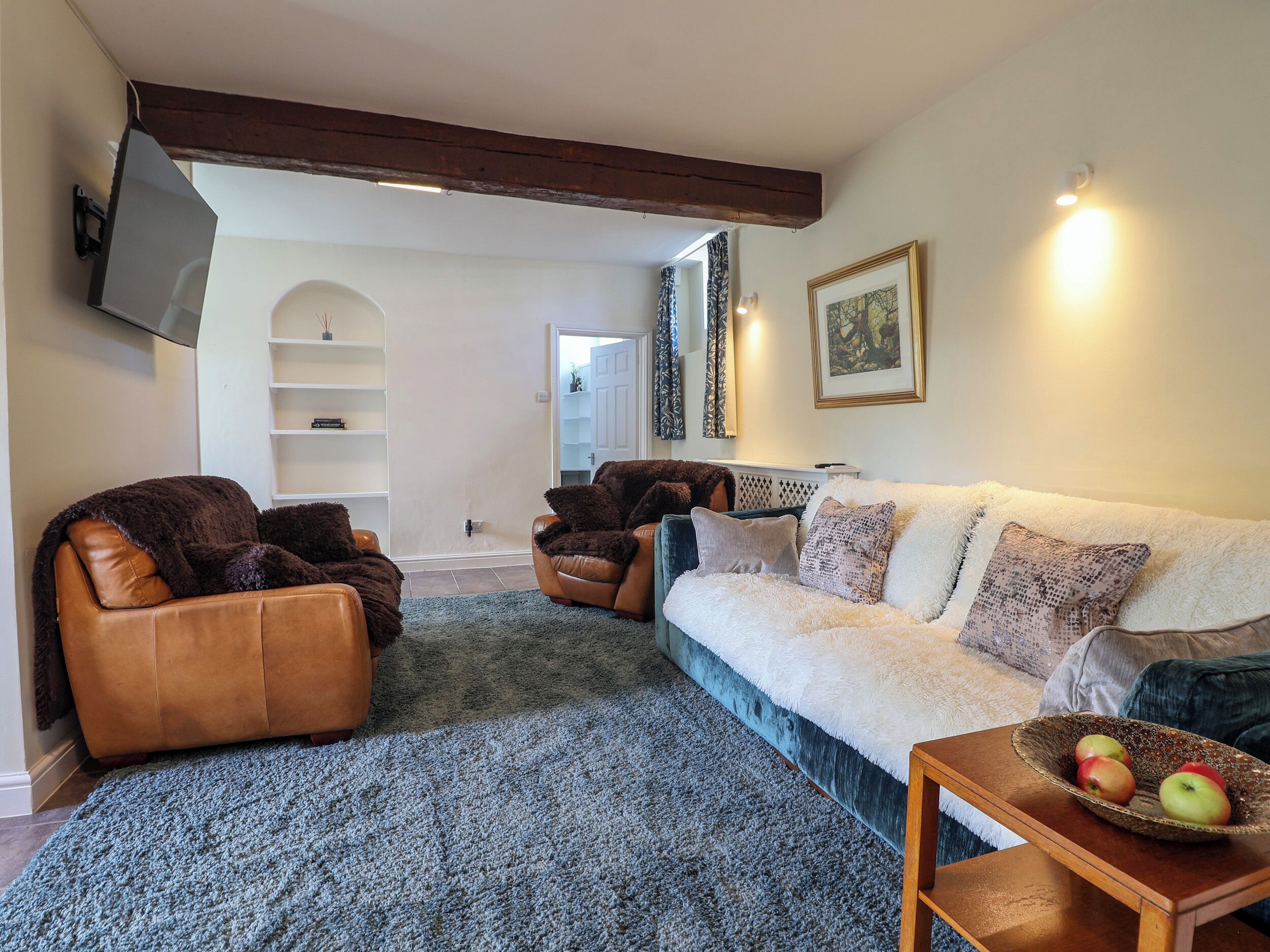 A living room with a sofa, two armchairs, a coffee table with fruit, and a TV mounted on the wall.