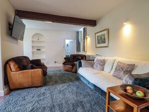 A living room with a sofa, two armchairs, a coffee table with fruit, and a TV mounted on the wall. - Linhay Barn (Saltash)