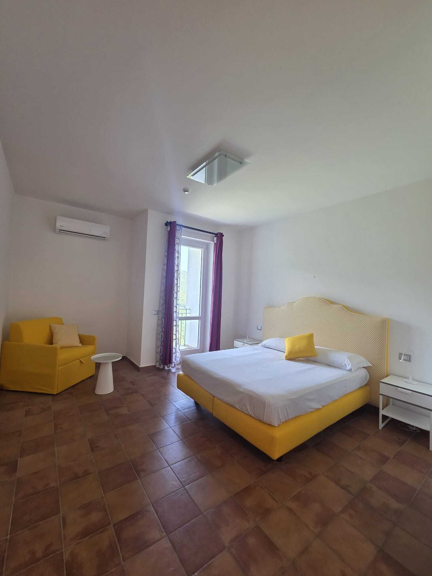 Un dormitorio con cama, una silla, una mesita, un equipo de aire acondicionado y una ventana con cortinas.