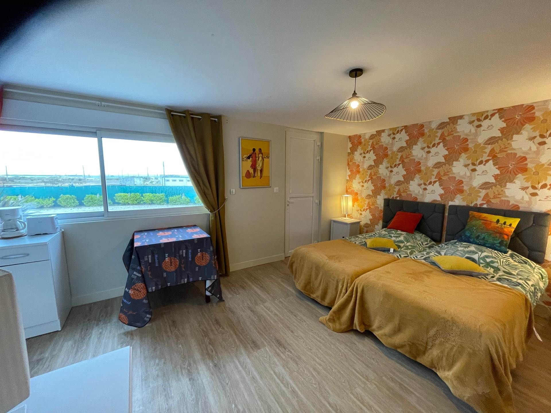 Une chambre à coucher comprenant un lit, un bureau, une chaise, une fenêtre avec des rideaux et un mur orné d’un motif floral.