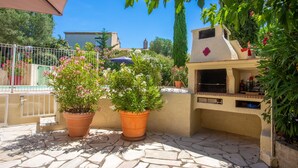 A patio with two terracotta planters, a barbecue grill, and a white building with a red flower on the wall. - La Thébaïde – 3-Star Villa with Private Pool near Fréjus-St-Raphaël (Puget-sur-Argens)