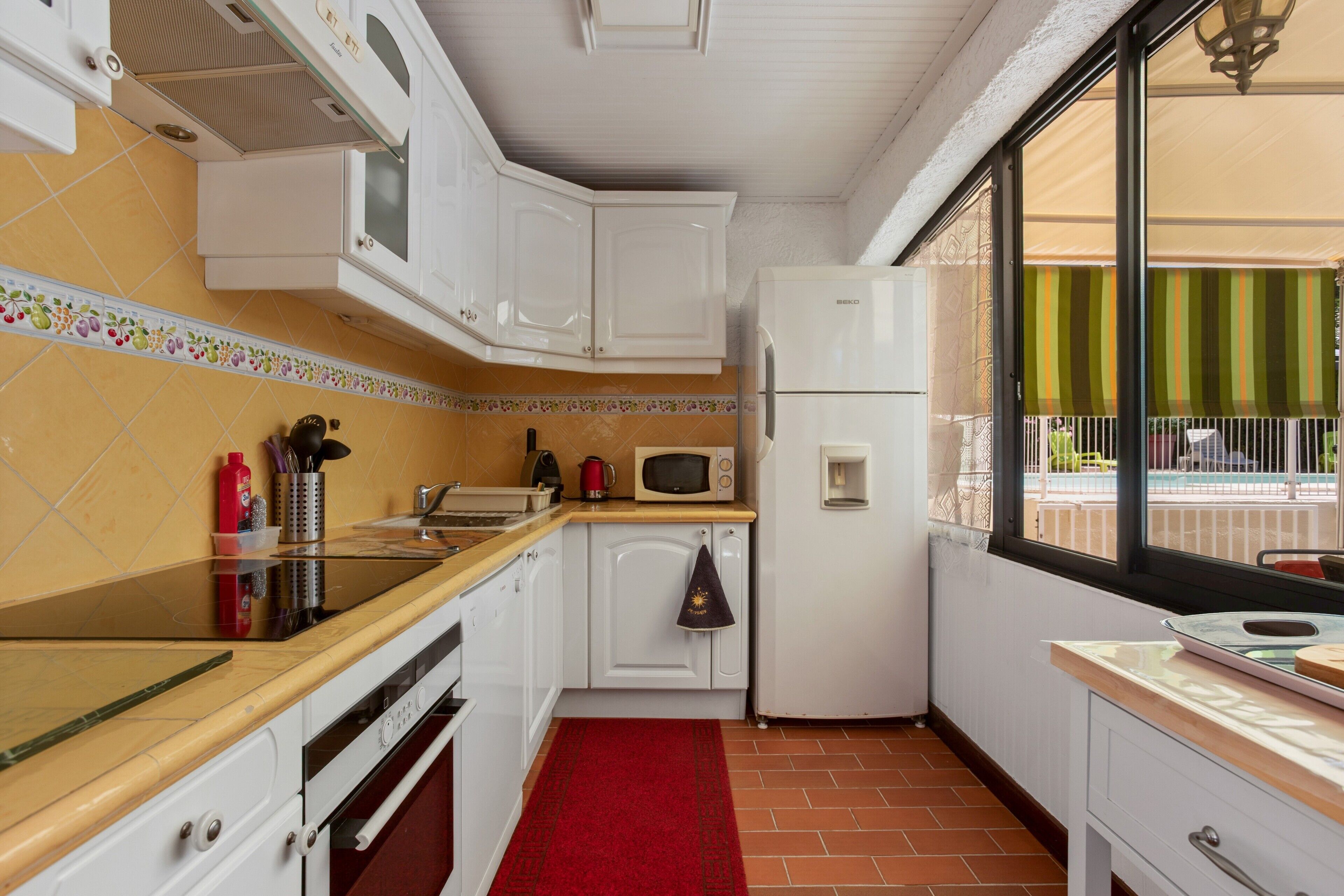Een compacte keuken met witte kastjes, een magnetron, een koelkast en een rood vloerkleed.