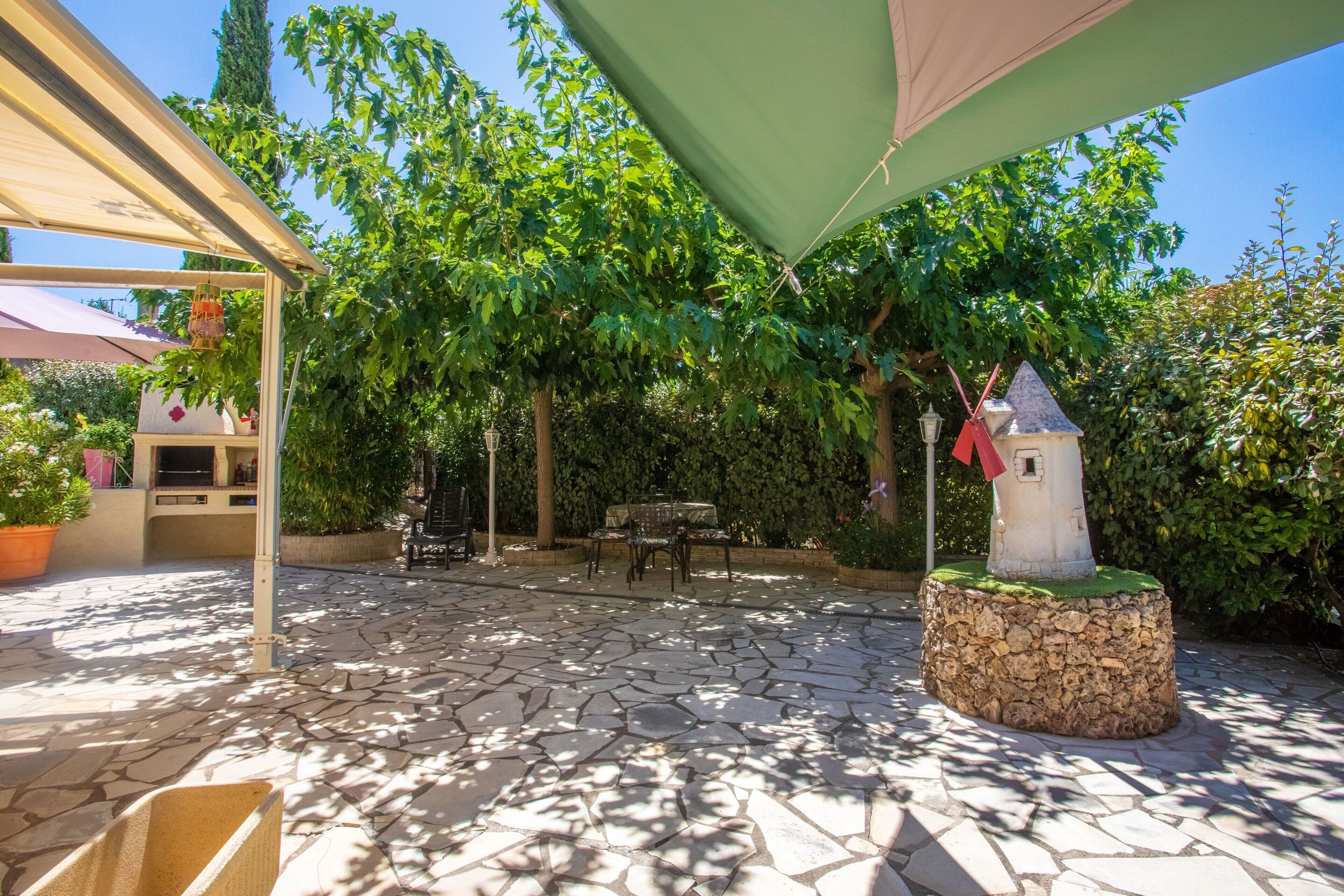 Une terrasse avec un sol en pierre, une pergola surmontée d’une tonnelle végétalisée, une petite structure blanche en forme de tour et une végétation luxuriante tout autour.