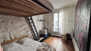 A bedroom with a loft bed, a ladder, a radiator, a window with curtains, and a large abstract painting. - A cozy 35 m² apartment near Montmartre (Paris)
