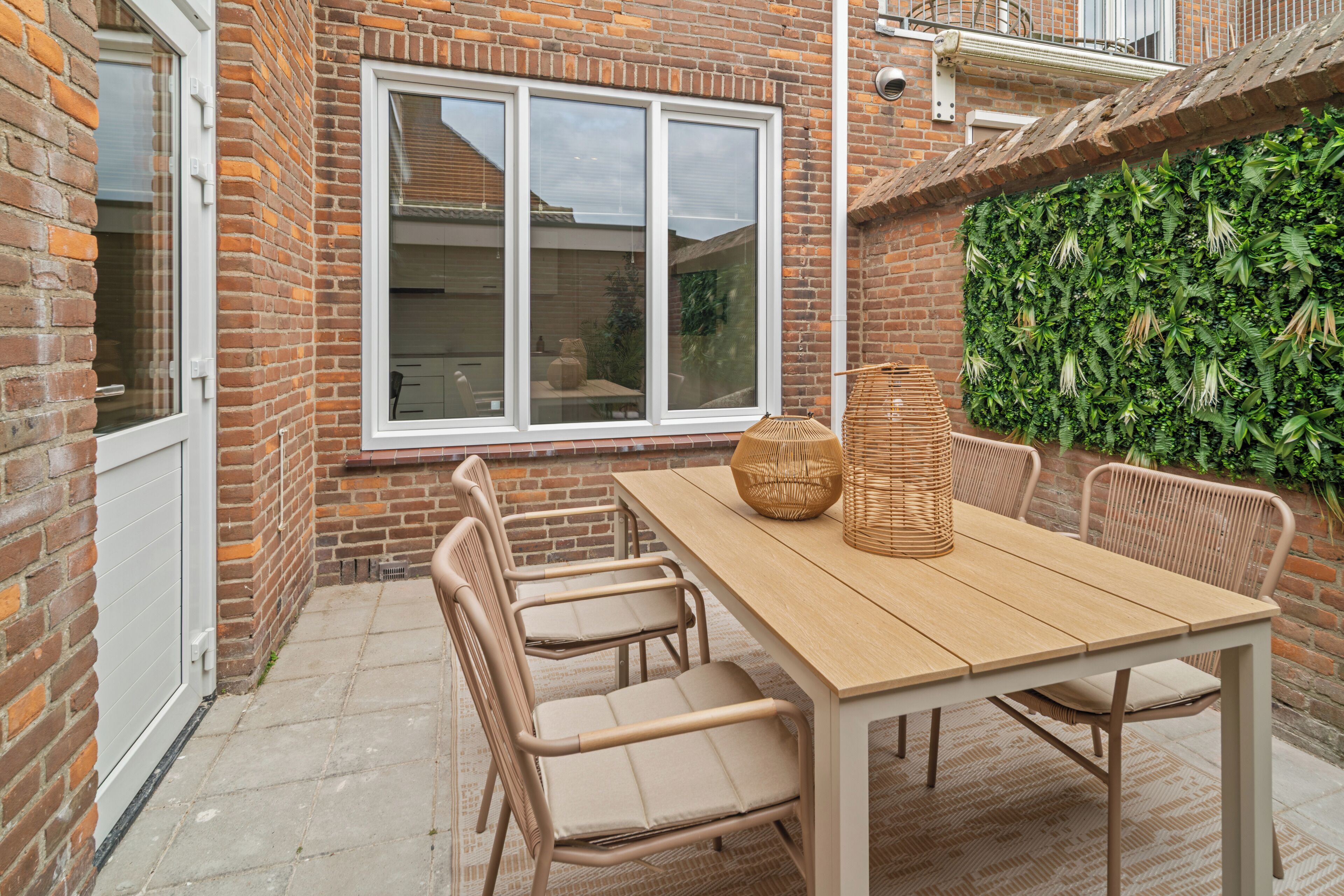 Un patio con una mesa y sillas de madera, una ventana de marco blanco y un muro de ladrillo.