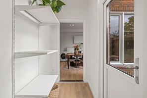 Un pasillo estrecho con estanterías blancas, un comedor con mesa y sillas, y una ventana que ofrece vistas a un muro de ladrillo y vegetación.