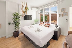 Quarto de hotel moderno com uma cama grande, televisão de tela plana, um vaso de planta e vista para o exterior através de uma janela ampla.