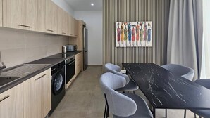 Eine moderne Küche mit Holzschränken, einer integrierten Waschmaschine, einem Essbereich mit schwarzem Tisch und grauen Stühlen sowie einem farbenfrohen abstrakten Gemälde an der Wand.