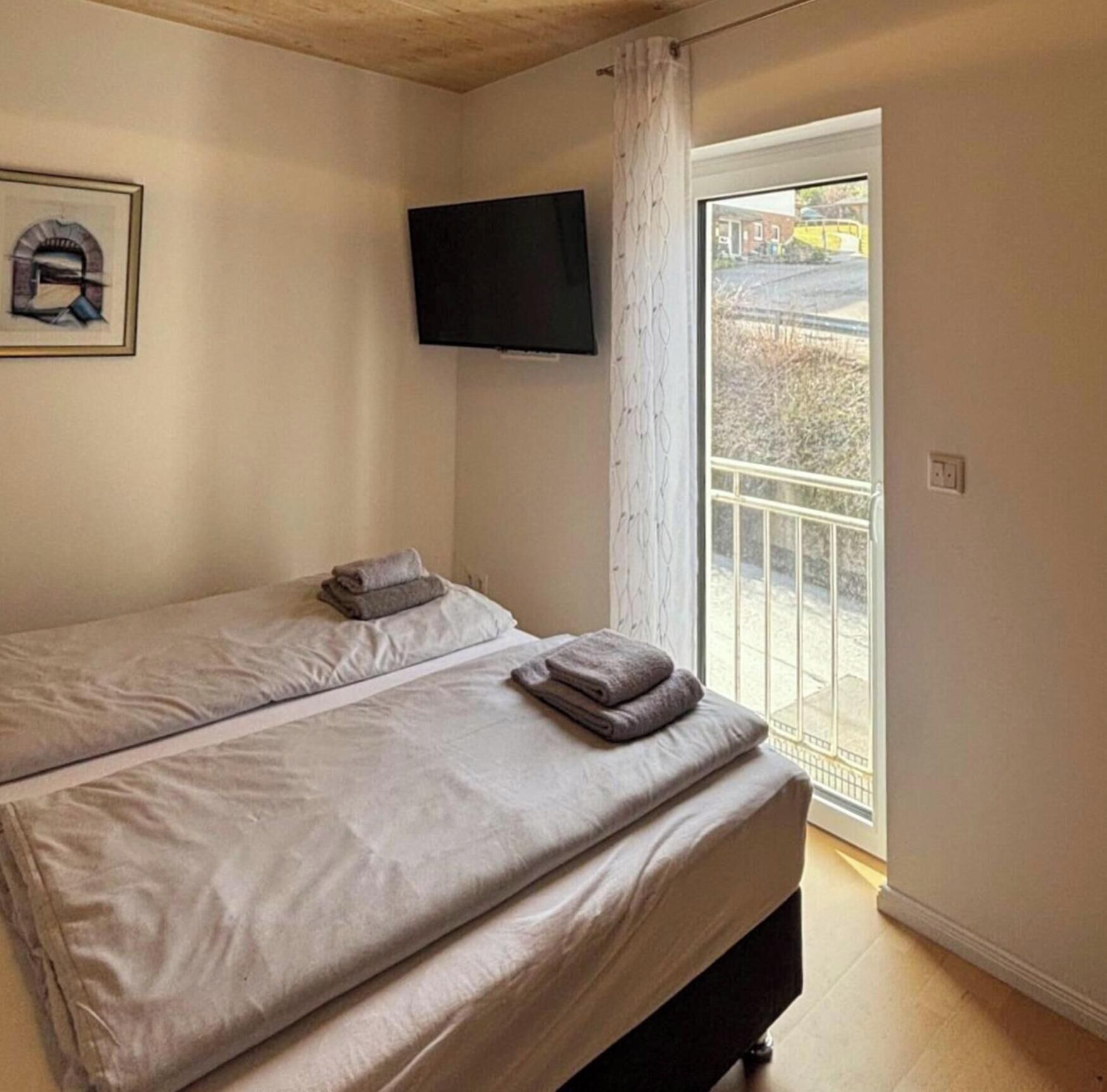 A bedroom with a bed, a TV mounted on the wall, and a window with a view of the outside.