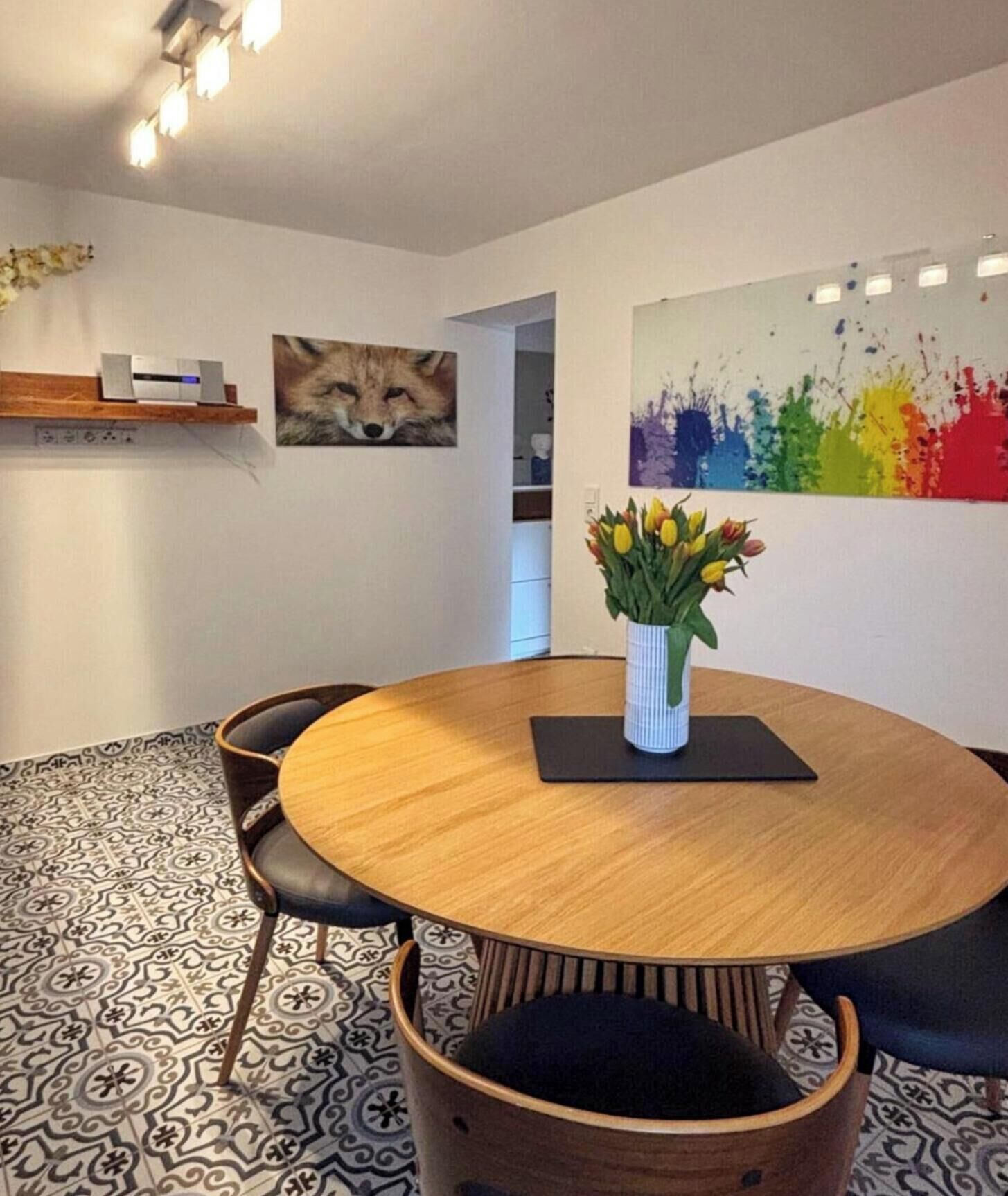 A round wooden table with a vase of flowers, surrounded by chairs, in a room with patterned flooring and artwork on the walls.