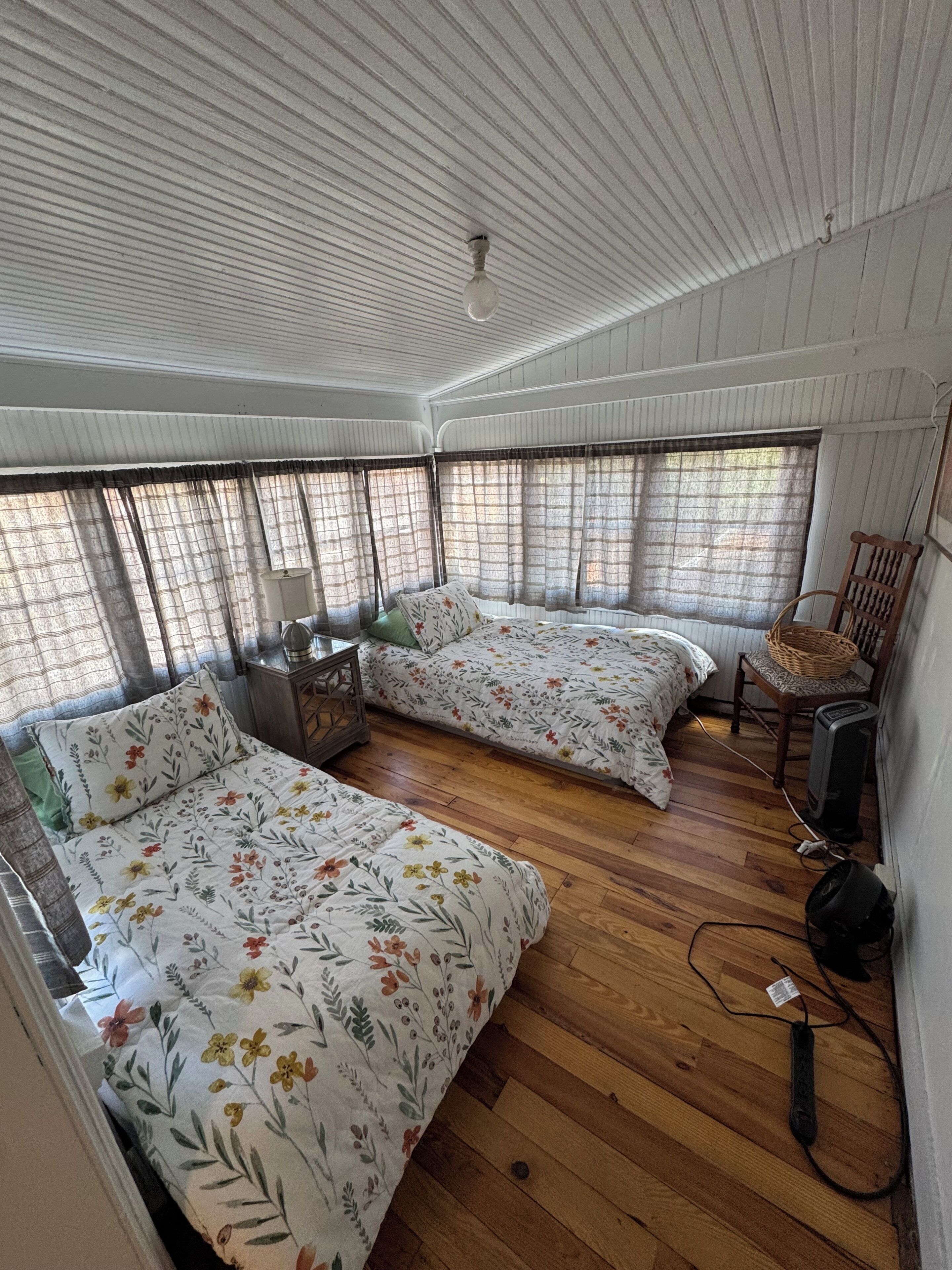 A room with two beds, a wooden floor, a chair, a lamp, and a fan.