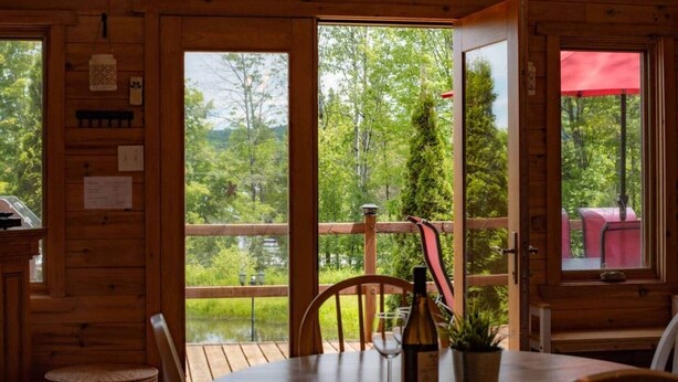 A wooden cabin with a dining area, a table with a bottle and glass, and a view of a forest and lake.