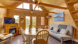 A wooden cabin interior with a dining table, chairs, a sofa, a fireplace, a television, and a painting of a landscape.