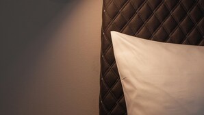 A bed with a quilted headboard and a white pillow, a wall-mounted light fixture, and a plain wall. - gambino hotel GOETHE26 (Munich)