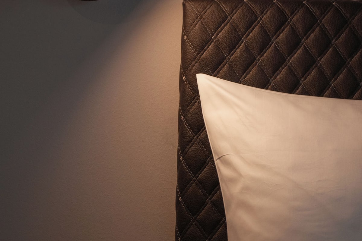 A bed with a quilted headboard and a white pillow, a wall-mounted light fixture, and a plain wall.