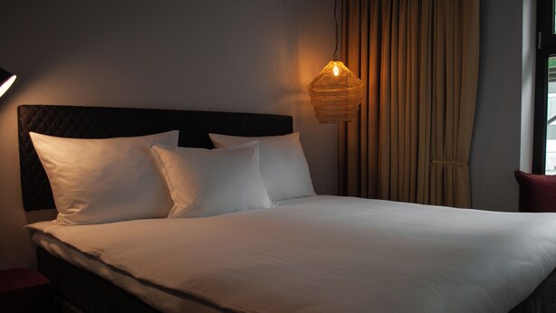 A neatly made bed with white linens and a dark headboard, a hanging light fixture, and a window with curtains. - gambino hotel GOETHE26 (Munich)