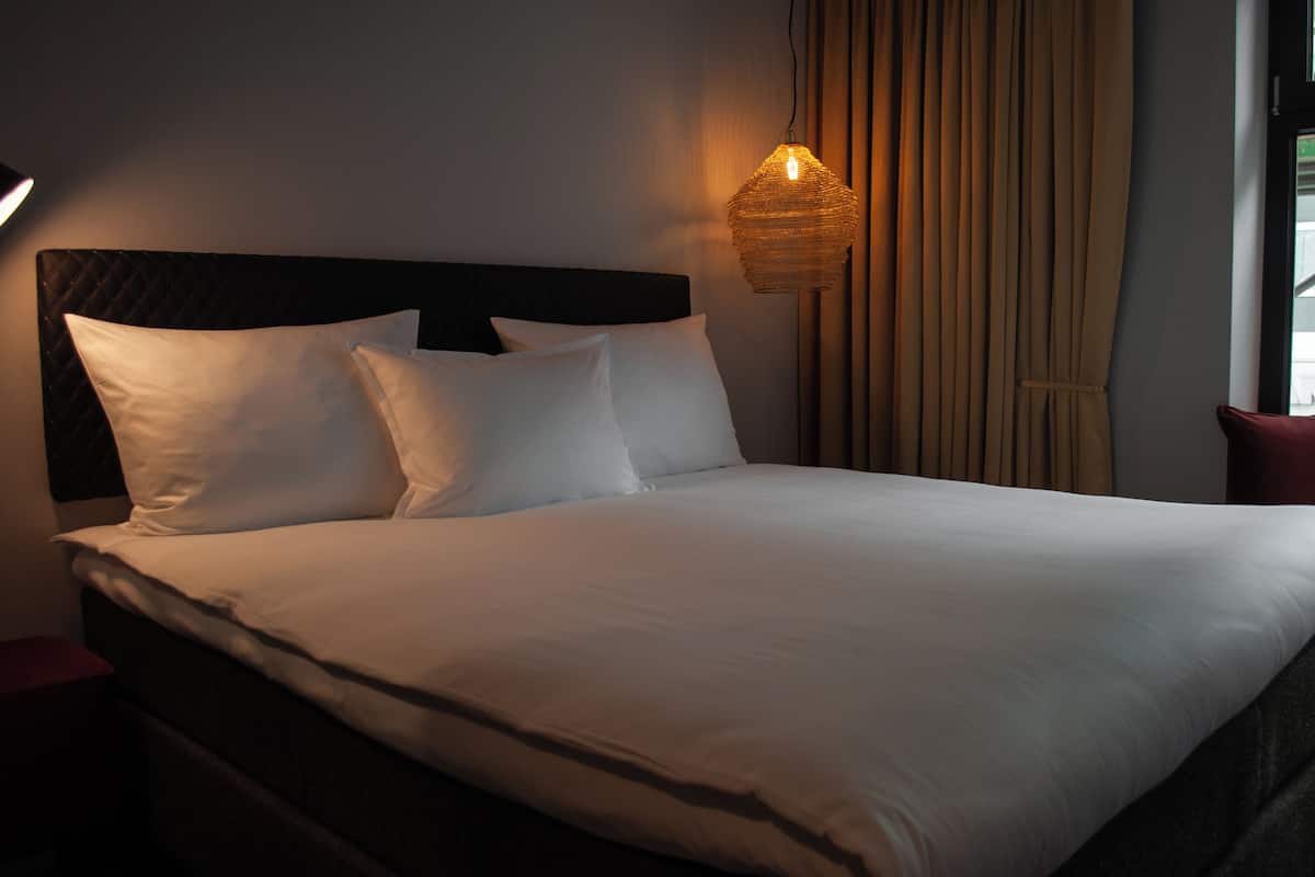 A neatly made bed with white linens and a dark headboard, a hanging light fixture, and a window with curtains.