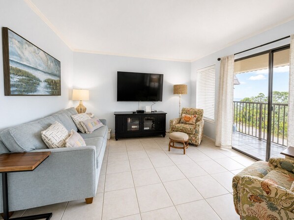 A living room with a sofa, a television, a side table, a chair, and a balcony with a view. - Ocean View - Admiral's Row 431- Professionally Managed by HHB&T Vacation Rentals (Hilton Head Island)