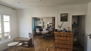 Quarto com cômoda de madeira, mesa pequena, banco, estante e porta que dá para um banheiro.