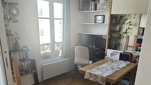Quarto com piso de madeira, uma mesa com toalha estampada, uma cadeira, um radiador e uma janela com cortinas.